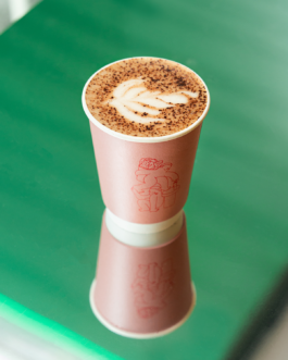 Coffee cup with Buddy Buddy x Oatly pop-up illustration and latte art on a green reflective surface.