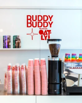 Coffee counter at the Buddy Buddy x Oatly pop-up in Printemps, Paris, with the collaboration logo displayed on the wall behind the espresso machine and branded cups stacked in front.