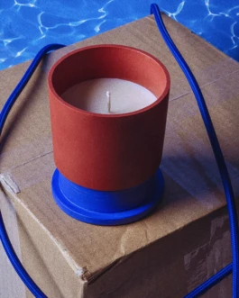 Hank Brussels Poly candle Son of a Beach in a red vessel with blue base, styled on a cardboard box with a marine-inspired rope and pool water reflections in the background.